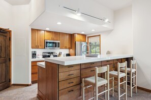 A modern kitchen with wooden cabinets, a large island with bar stools, stainless steel appliances, and a window providing natural light. - Slopeside Luxury, Pool, Hot Tubs, Conceirge | LB114 (Beaver Creek)