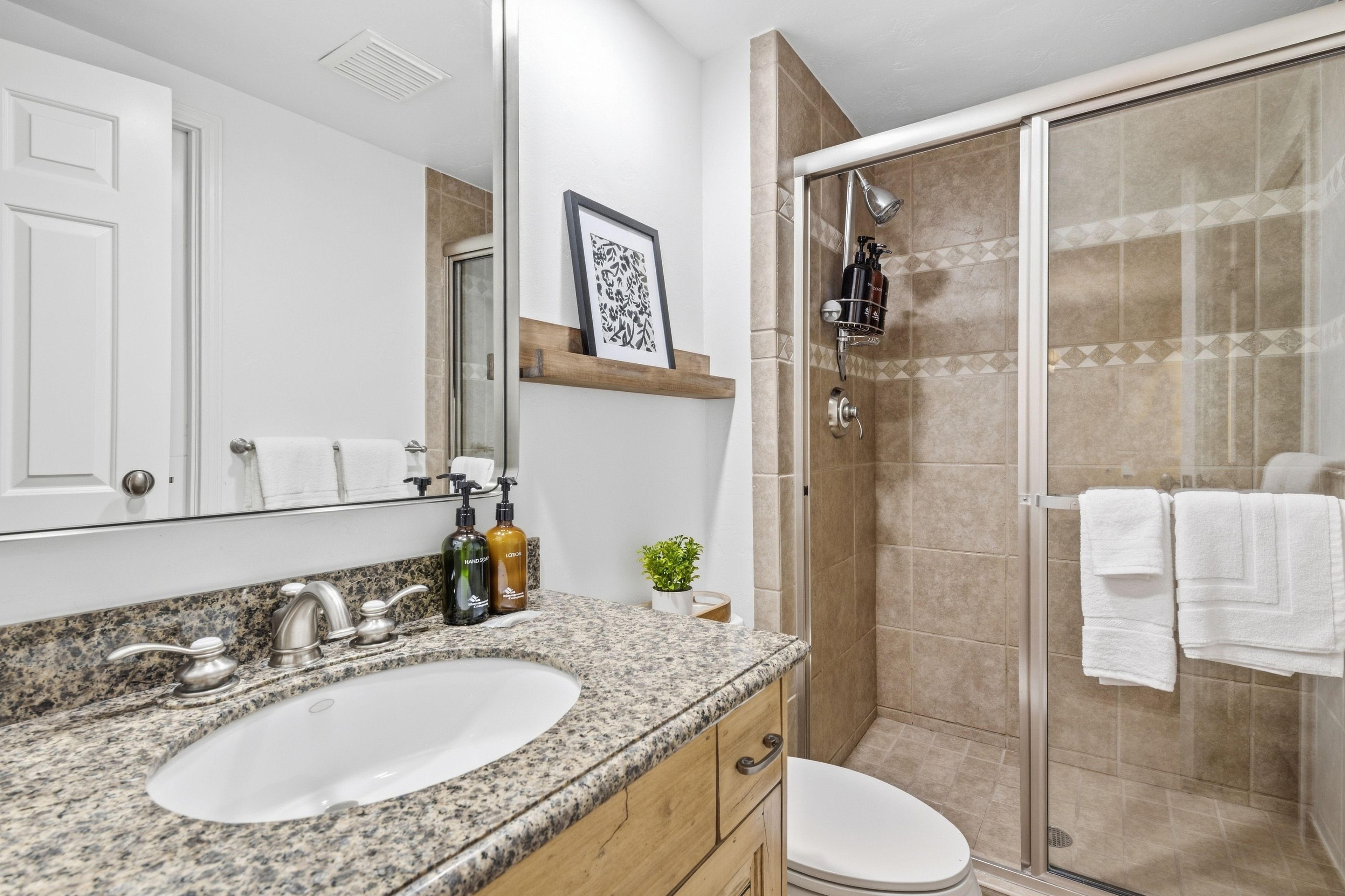Un baño con lavamanos de encimera de granito, un espejo, un cuadro enmarcado, una ducha con puerta de cristal y un perchero.