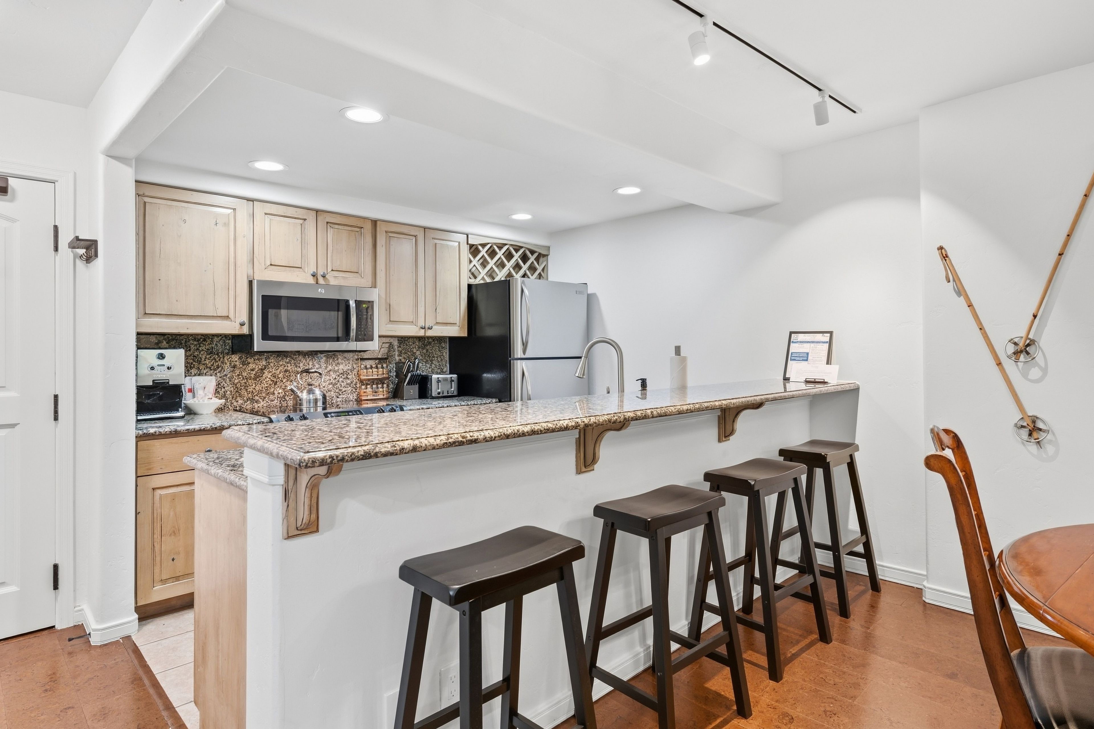 A modern kitchen with granite countertops, stainless steel appliances, and wooden cabinets. There are four stools at the bar area.