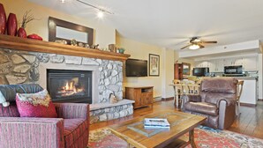 Una sala de estar con chimenea de piedra, un televisor montado encima y un comedor con una cocina al fondo.