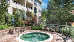 Zona de piscina con sillas y jacuzzi, rodeada de árboles y un edificio con balcones.
