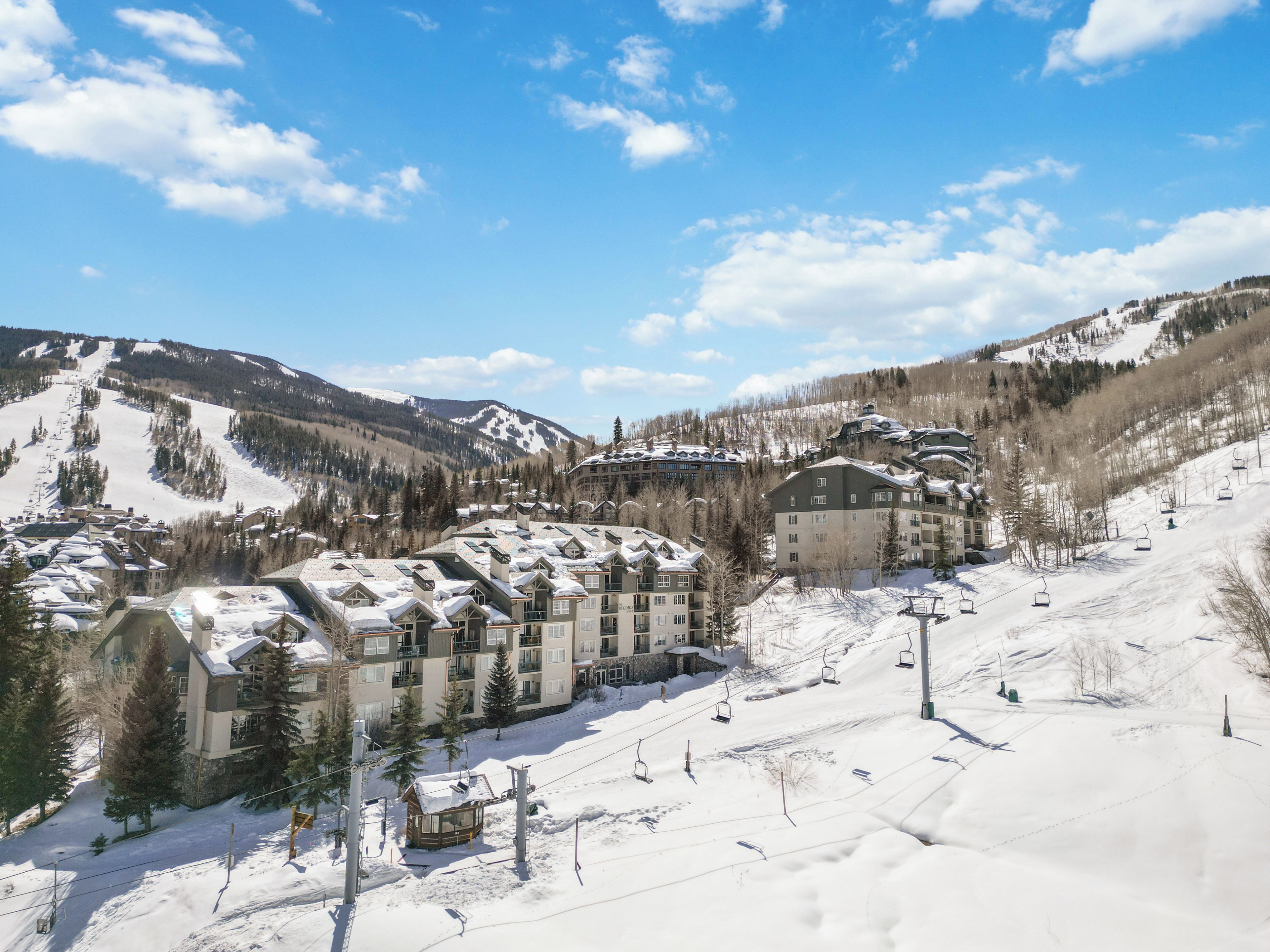 A snow-covered ski resort with multiple buildings, including a large hotel with multiple floors and balconies, surrounded by trees and mountains.