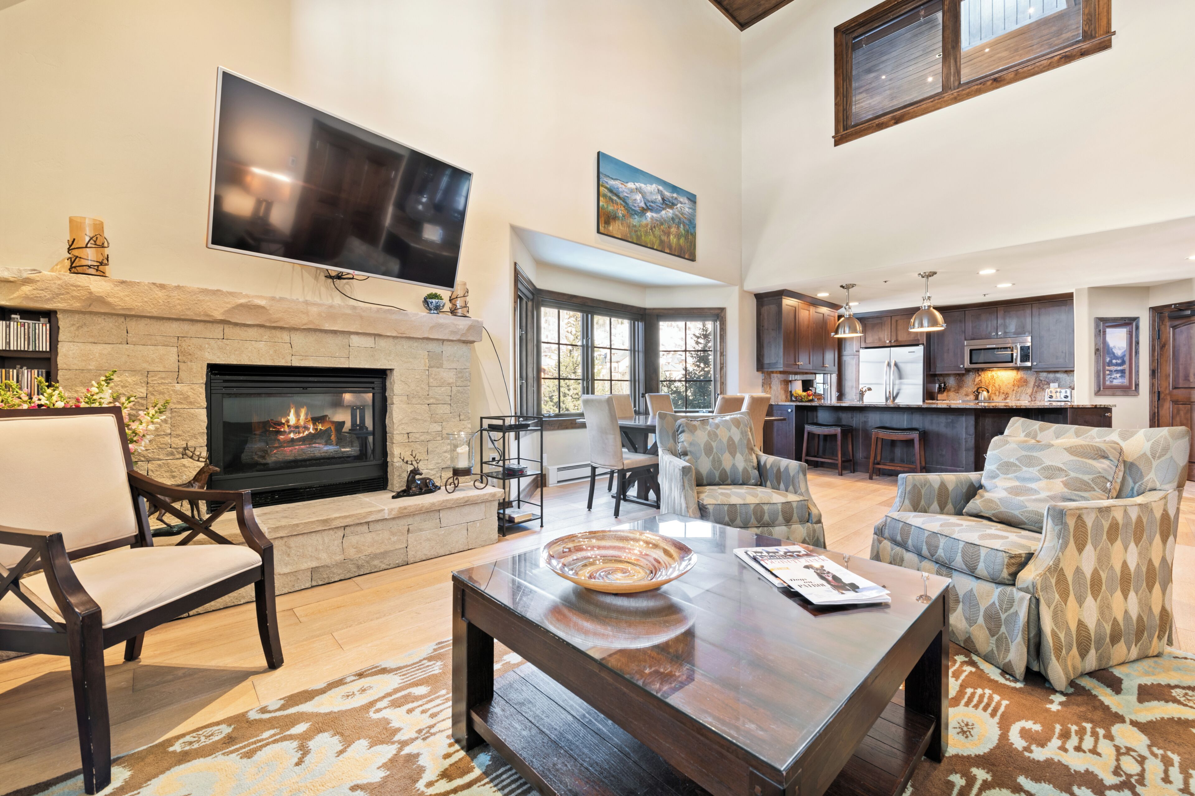 A spacious living area with a fireplace, comfortable seating, a flat-screen TV, and a kitchen in the background.