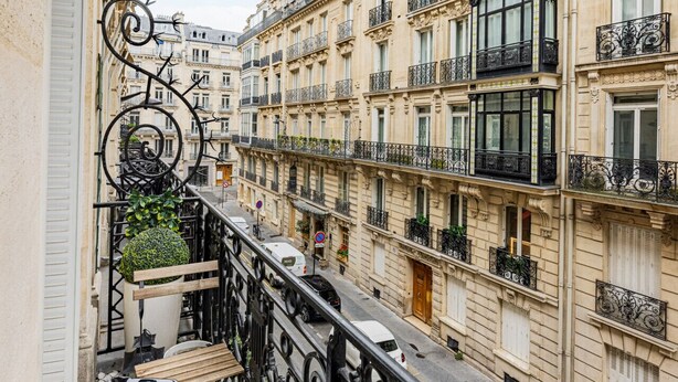 A narrow street with buildings on either side, balconies with black wrought iron railings, and a table with chairs on the left balcony. - Champs Elysées-Avenue Montaigne-2 rooms (Paris)