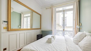 A hotel room with a large mirror, a bed with white linens, a radiator, and a view of buildings through a window. - Champs Elysées-Avenue Montaigne-2 rooms (Paris)