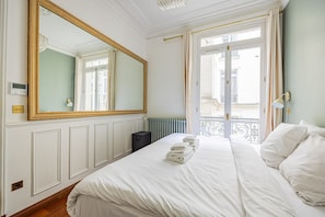 Ein Hotelzimmer mit einem großen Spiegel, einem Bett mit weißen Leinen, einem Heizkörper und Blick auf Gebäude durch ein Fenster.