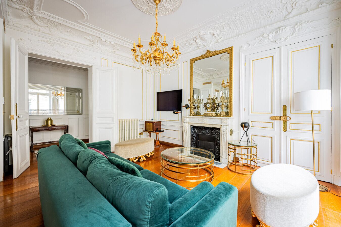 Un salon spacieux avec un canapé vert, un pouf blanc, une table basse en verre, une cheminée et un grand miroir.