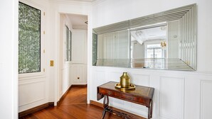 A hallway with a wooden floor, a large mirror, a brass vase on a wooden console table, and a door with a glass panel. - Champs Elysées-Avenue Montaigne-2 rooms (Paris)