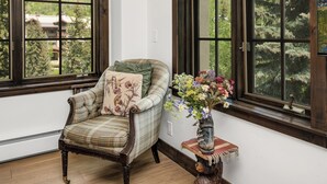Ein Zimmer mit einem kariertem Sessel, einer Blumenvase und einem Fenster mit Blick auf Bäume und Gebäude.