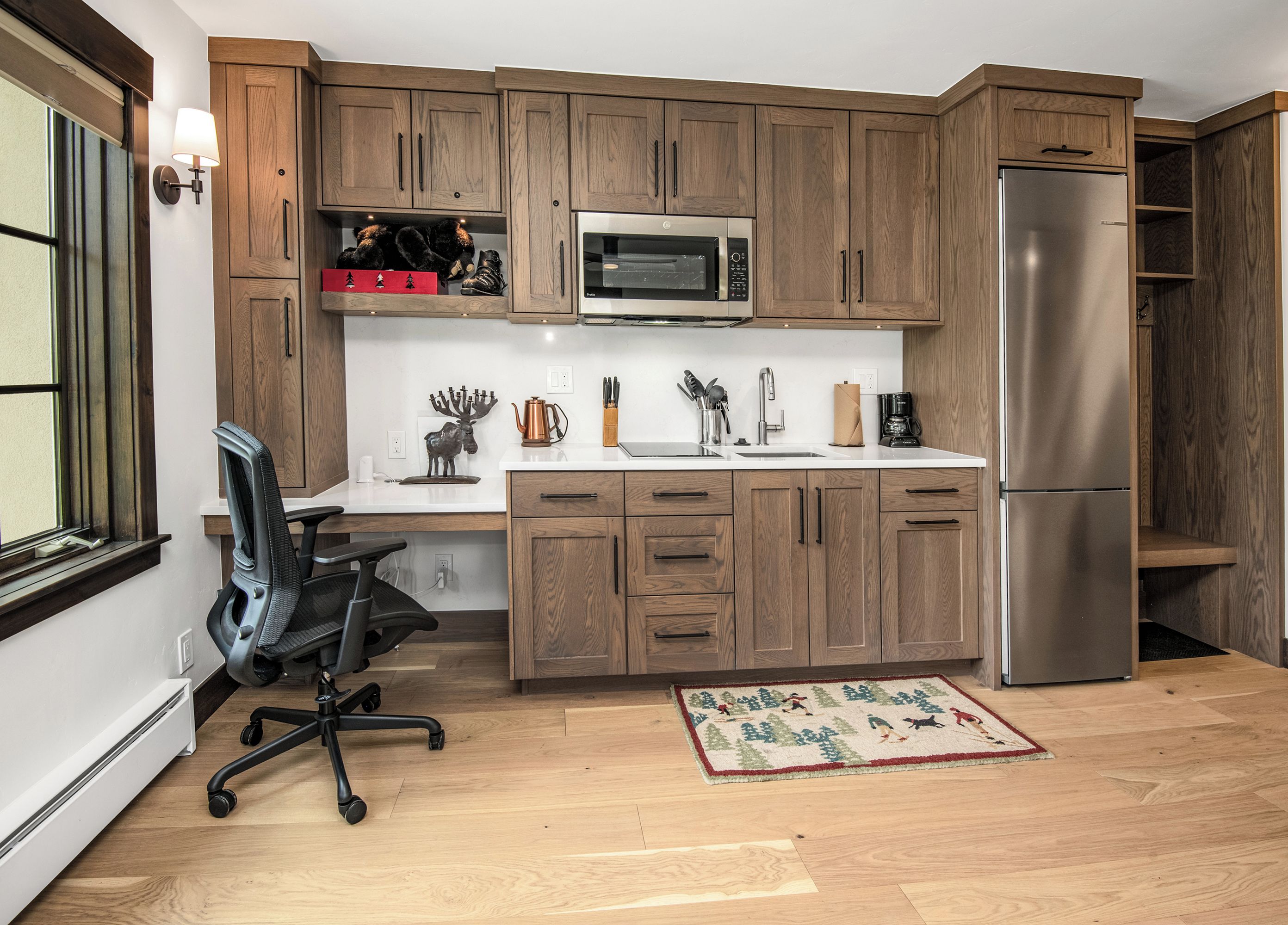 A kitchen with wooden cabinets, a stainless steel refrigerator, a microwave, a coffee maker, and a rug with a Christmas theme.