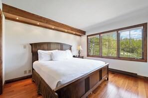 Ein Schlafzimmer mit einem großen Bett, einem Holzkopfstück, einem Nachttisch mit einer Lampe und einem großen Fenster mit Blick auf Bäume.