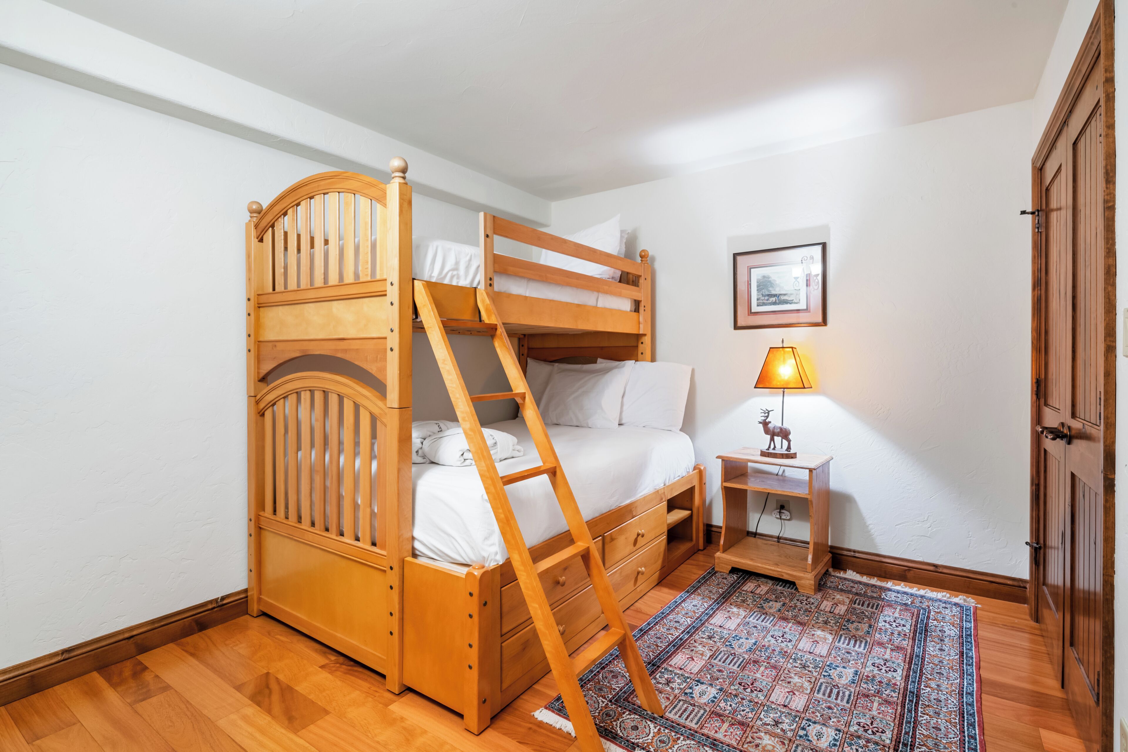 Une chambre avec un lit superposé en bois, une petite table de chevet avec une lampe, un tapis à motifs et une porte.