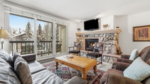 Una sala de estar con chimenea de piedra, televisor y balcón con vistas a árboles nevados.