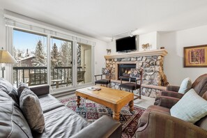 Ein Wohnzimmer mit Kamin aus Stein, einem Fernseher und einem Balkon mit Blick auf schneebedeckte Bäume.