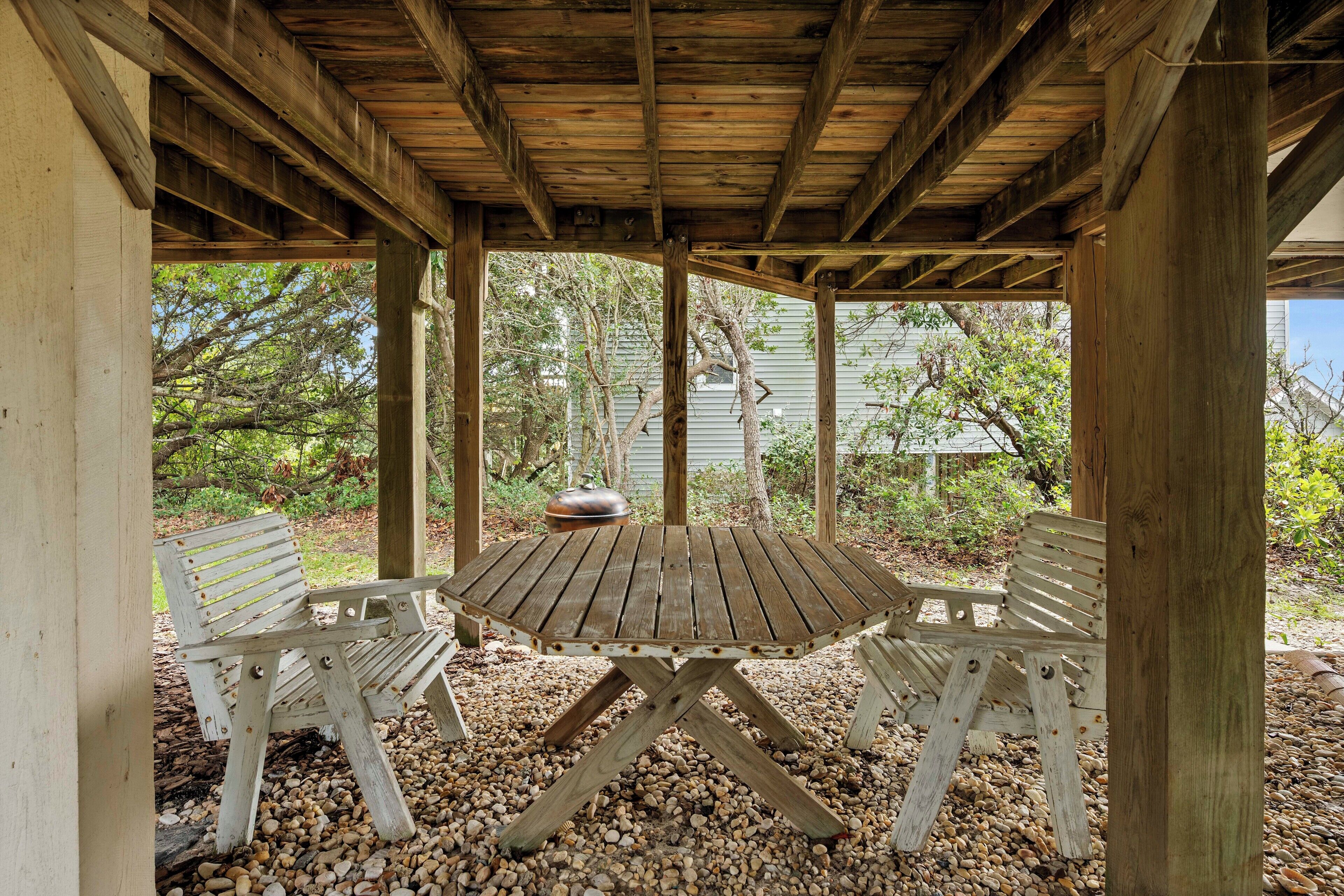 A wooden pergola with a table and two chairs, surrounded by pebbles and trees.