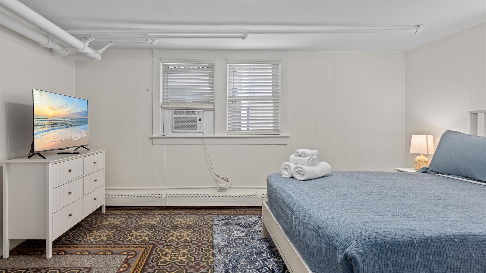 Ein Schlafzimmer mit einem Bett, einem Fernseher auf einer Kommode, einem Fenster mit Jalousien und einem gemusterten Teppich.