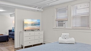 Ein Hotelzimmer mit Fernseher auf einer Kommode, einem Bett, einem Fenster mit Jalousien und einer Wand-Klimaanlage.