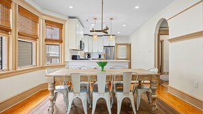 A dining area with a wooden table, white chairs, a green centerpiece, and a kitchen in the background.