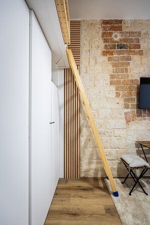 A room with a wooden loft bed, a brick wall, a white wardrobe, and a small seating area with a cushion. - Batignolles - Cosy renovated studio apartment (Paris)