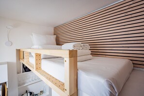 A bunk bed with white bedding, a wooden slat headboard, and a shelf with glasses and bottles. - Batignolles - Cosy renovated studio apartment (Paris)