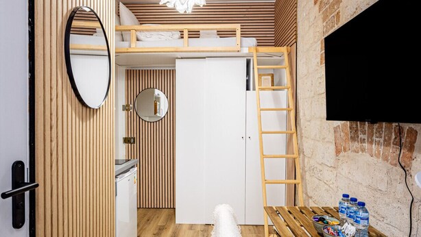 Une pièce compacte avec un lit en mezzanine, une table à manger en bois, une chaise blanche, un luminaire suspendu, un miroir et un téléviseur fixé sur un mur en briques.