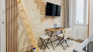 Une pièce avec un mur en briques, un plancher en bois, un téléviseur fixé au mur, une table avec des chaises et une fenêtre avec des rideaux.