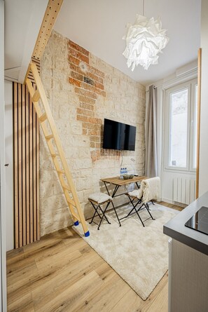 A room with a brick wall, wooden floor, a TV mounted on the wall, a table with chairs, and a window with curtains. - Batignolles - Cosy renovated studio apartment (Paris)