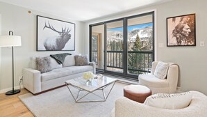 Una sala de estar moderna con sofá, mesa de centro y sillón, ventanas amplias con vista a la montaña, y obras de arte en las paredes.