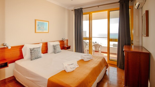 A hotel room with a bed, a wooden headboard, a nightstand, a desk, a chair, a balcony with a view, and a painting on the wall. - FLH Monte Gordo Balcony Flat (Monte Gordo)