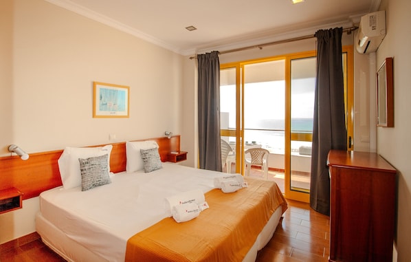 A hotel room with a bed, a wooden headboard, a nightstand, a desk, a chair, a balcony with a view, and a painting on the wall. - FLH Monte Gordo Balcony Flat (Monte Gordo)