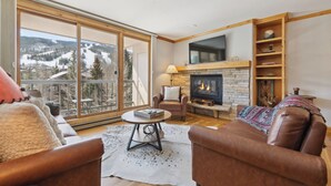 Ein Wohnzimmer mit Kamin, einer Couch, einem Sessel, einem Fernseher und einem großen Fenster mit Blick auf eine verschneite Landschaft.