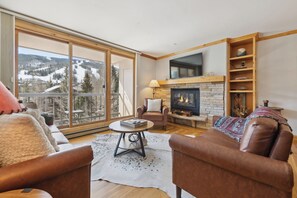 Ein Wohnzimmer mit Kamin, einer Couch, einem Sessel, einem Fernseher und einem großen Fenster mit Blick auf eine verschneite Landschaft.