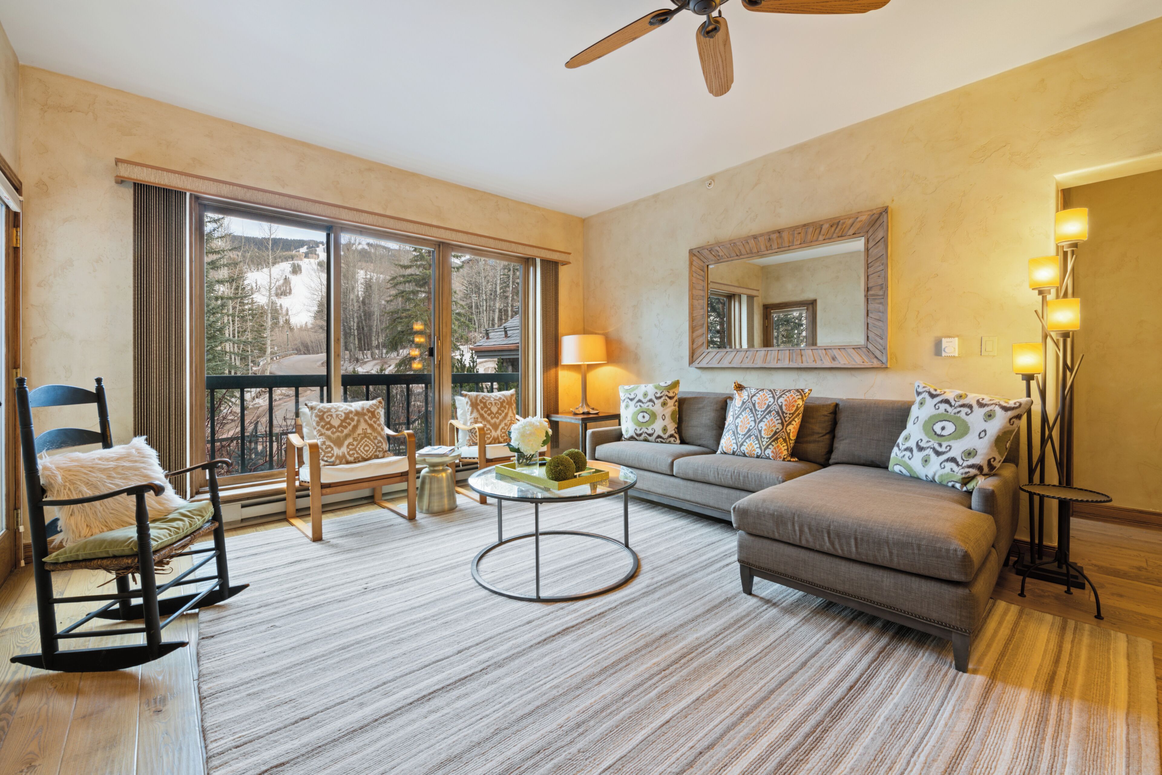 A spacious living room with a large window, a ceiling fan, a sofa, a coffee table, and a rocking chair.