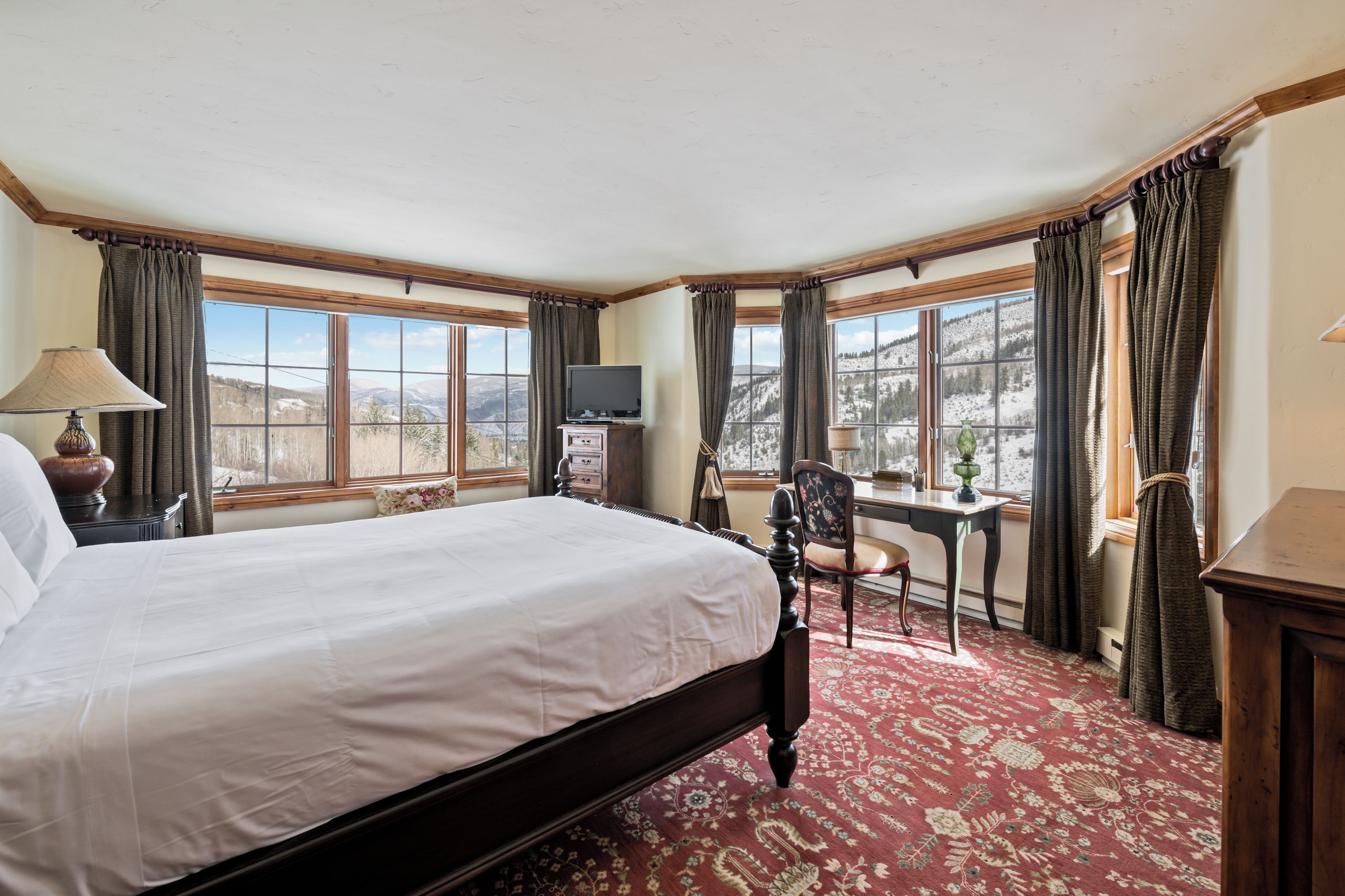 A bedroom with a large bed, a desk, a chair, a television, and a window with curtains.