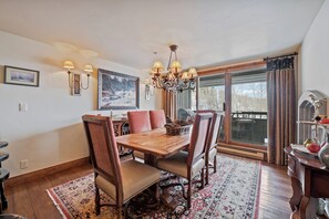 A dining area with a wooden table, chairs, a chandelier, wall sconces, and a large window with curtains. - Beaver Creek Ski-In/Ski-Out w/Mountain View| UB407 (Beaver Creek)