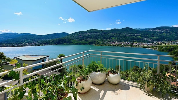 A balcony with a view of a lake, mountains, and a town in the background. - Luxury Lakeview Penthouse (Montagnola)
