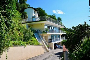 A two-story building with a balcony on the upper level, a staircase leading to the entrance, and a car parked in the driveway. - Luxury Lakeview Penthouse (Montagnola)