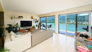 A modern living room with a large TV, a sofa, and a balcony with a scenic view. - Luxury Lakeview Penthouse (Montagnola)