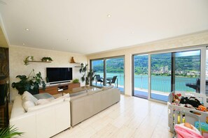 A modern living room with a large TV, a sofa, and a balcony with a scenic view. - Luxury Lakeview Penthouse (Montagnola)