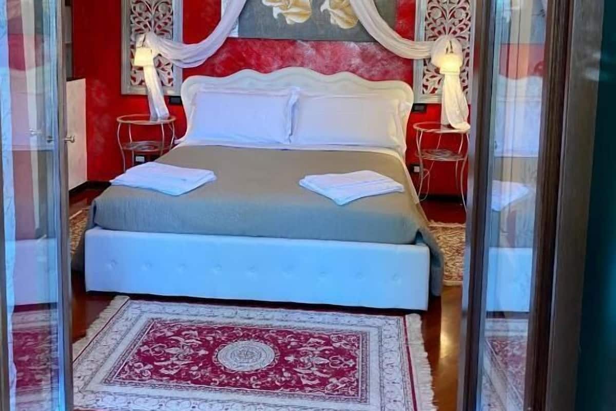 A bedroom with a bed, chandelier, red walls, and a patterned rug.
