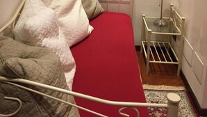 Un dormitorio con una colcha roja, estructura de cama de metal blanco, una mesita de noche con lámpara, un cuadro enmarcado en la pared y un radiador.