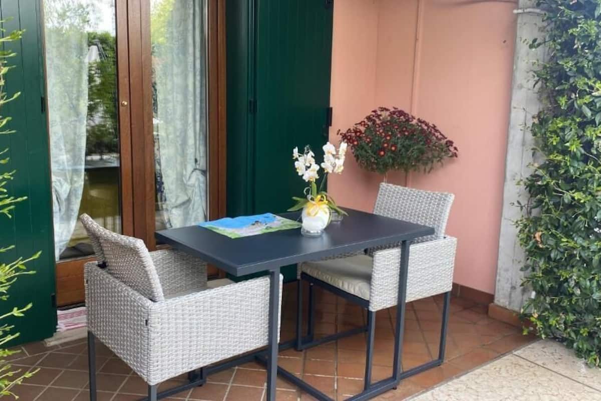 A patio with wicker chairs, a table, a vase with flowers, and a view through a glass door.