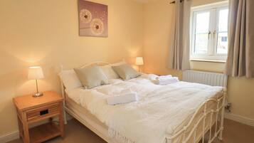 A neatly made bed with white linens, a bedside table with a lamp, and a window with curtains.