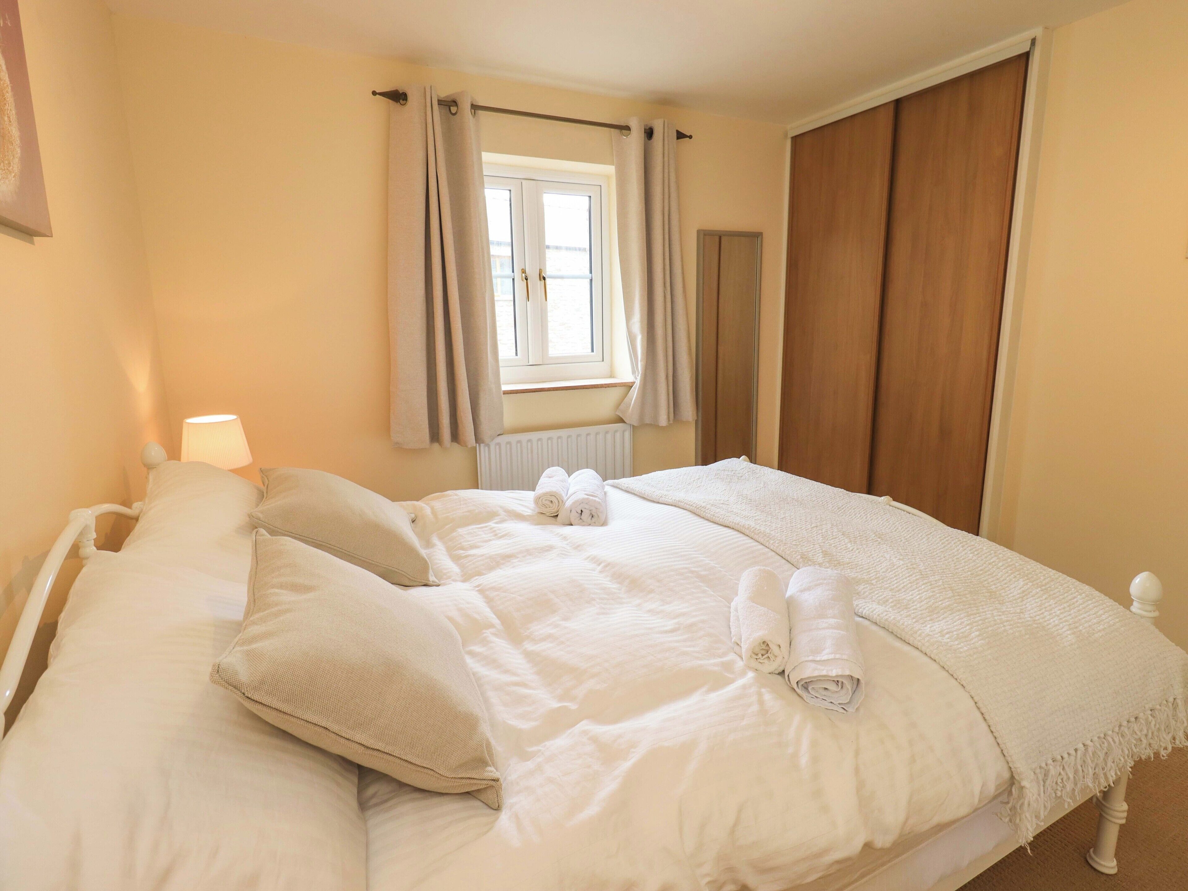 A neatly made bed with pillows and rolled towels, a window with curtains, a radiator, and a wooden wardrobe.