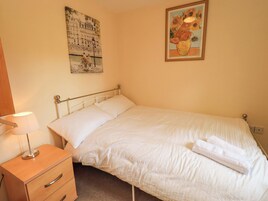 Una cama individual con ropa blanca de cama, una mesita de noche con lámpara, una mesita auxiliar de madera y dos obras de arte colgadas en la pared.