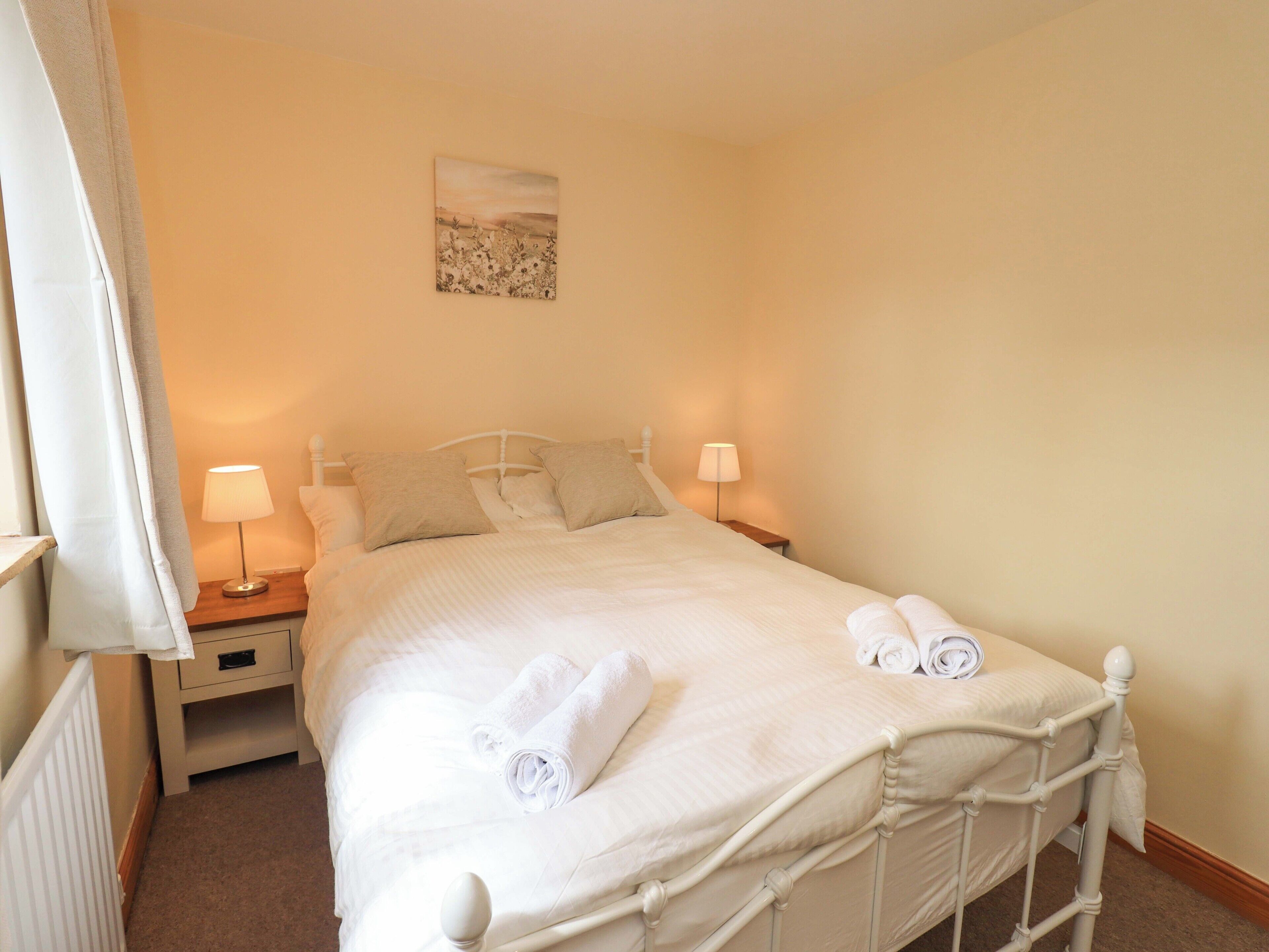 A neatly made bed with white linens and towels, two bedside lamps, a nightstand, and a painting on the wall.