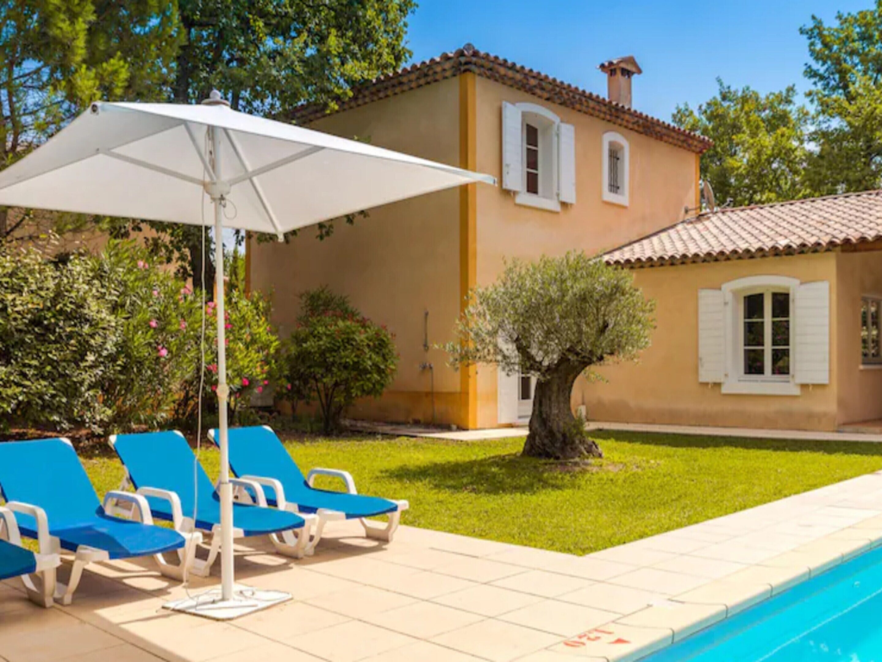 Une maison avec une piscine, des fauteuils de jardin bleus et un parasol blanc.