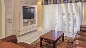 Una sala de estar con televisor, ventilador de techo, chimenea y un ventanal con persianas.