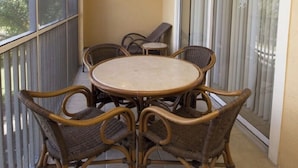Un balcón con una mesa redonda y sillas de mimbre, una mesita con una silla, y un ventanal corredera.