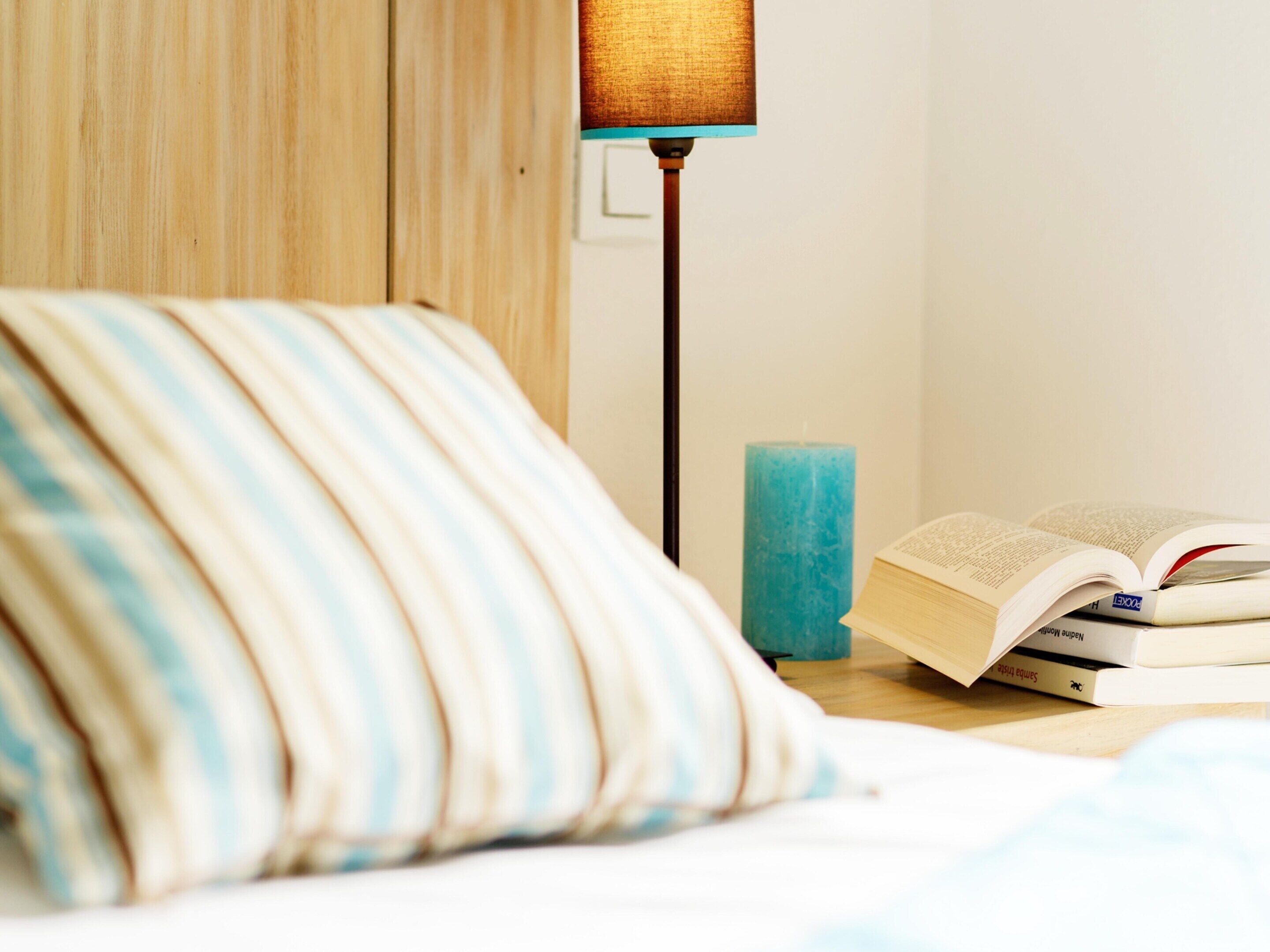 A neatly made bed with a striped pillow, a teal candle, and a book on a bedside table.